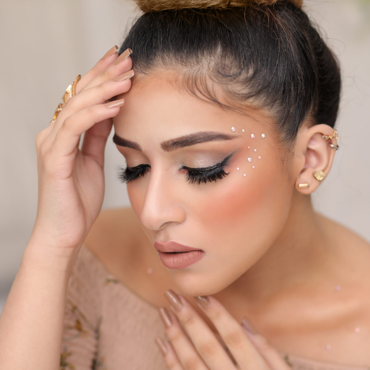 Close-up portrait of a woman with glowing skin, nude-toned makeup, and a sleek top bun hairstyle.