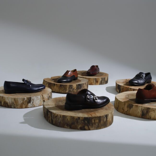 Premium leather dress shoes in black, brown, and burgundy displayed on round wooden platforms under studio lighting